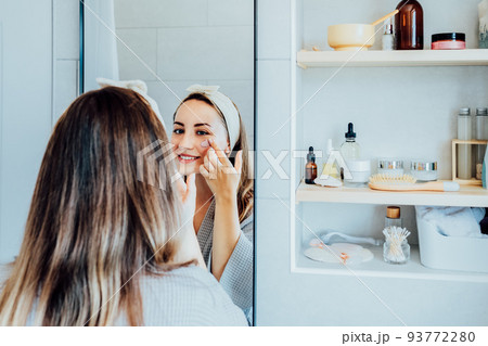 Young woman in bathrobe looking in the mirror and applying facial natural cosmetic clay mask on her face in bathroom. Cosmetic procedures for skin care at home. Beauty self-care. Selective focus. Young woman in bathrobe looking in the mirror and applying facial natural cosmetic clay mask on her face in bathroom. Cosmetic procedures for skin care at home. Beauty self-care. Selective focus. 93772280