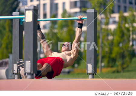 Young strong muscular man doing strength exercises on workout ground at public park in summer morning. Sport, fitness, healthy lifestyle concept Young strong muscular man doing strength exercises on workout ground at public park in summer morning. Sport, fitness, healthy lifestyle concept 93772529