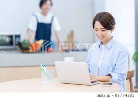 リビングでテレワークをするミドル女性とキッチンで料理をする男性 93774235