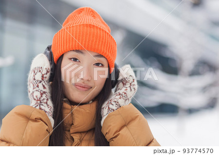 Close-up portrait of a young beautiful Asian woman looking at the camera and smiling while listening to music with big headphones, dressed warmly, on a snowy winter day Close-up portrait of a young beautiful Asian woman looking at the camera and smiling while listening to music with big headphones, dressed warmly, on a snowy winter day 93774570