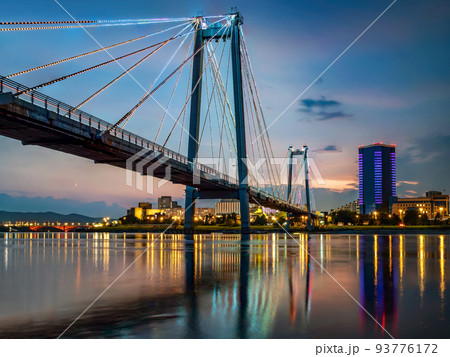 Suspension bridge bridge across the river at night. Night illumination of buildings, reflections. Suspension bridge bridge across the river at night. Night illumination of buildings, reflections. 93776172