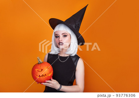 Beautiful sexy woman in witch hat and costume holding carved pumpkin on orange background. 93776306