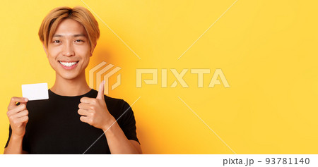 Close-up of satisfied asian guy recommend bank, showing credit card and standing over yellow background 93781140