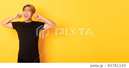 Portrait of annoyed and bothered asian guy grimacing, shut ears disturbed by loud noise, standing yellow background 93781150
