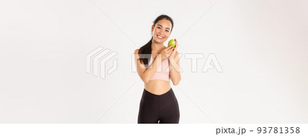 Sport, wellbeing and active lifestyle concept. Smiling cute asian fitness coach, girl athlete in sportswear showing green apple, sit on diet and workout to gain perfect body, white background 93781358