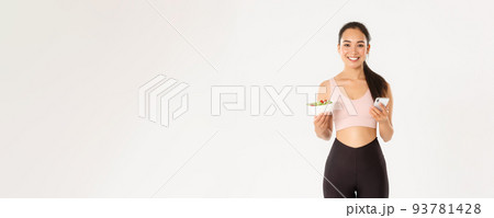 Sport, wellbeing and active lifestyle concept. Smiling slim asian fitness girl in sportswear, holding salad and mobile phone, using eating reminder app, diet control application, checking calories Sport, wellbeing and active lifestyle concept. Smiling slim asian fitness girl in sportswear, holding salad and mobile phone, using eating reminder app, diet control application, checking calories 93781428