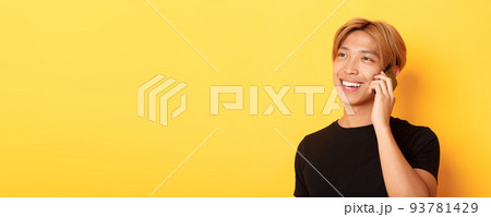 Close-up of handsome carefree asian guy with fair hair, talking on mobile phone and smiling happy, standing yellow background Close-up of handsome carefree asian guy with fair hair, talking on mobile phone and smiling happy, standing yellow background 93781429