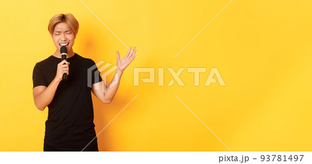 Handsome asian male singer, korean guy singing song at karaoke in microphone with passion, standing over yellow background Handsome asian male singer, korean guy singing song at karaoke in microphone with passion, standing over yellow background 93781497