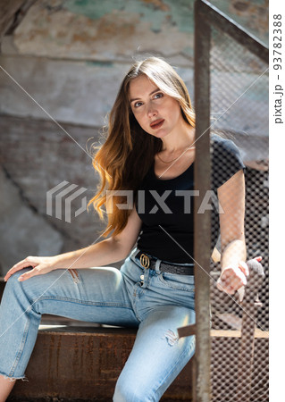 Woman in black t-shirt and jeans standing near wire fencing in abandoned factory Woman in black t-shirt and jeans standing near wire fencing in abandoned factory 93782388
