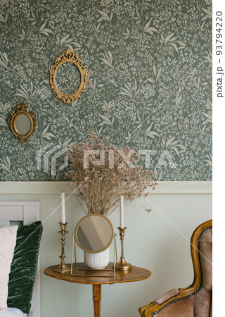 Dried flowers in a vase, candles on a classic candlestick and a table mirror stand on the wooden bedside table by the bed Dried flowers in a vase, candles on a classic candlestick and a table mirror stand on the wooden bedside table by the bed 93794220