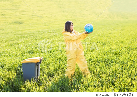 A woman in a yellow protective suit stands in the middle of a green field and holds a globe in her hands, next to a garbage can A woman in a yellow protective suit stands in the middle of a green field and holds a globe in her hands, next to a garbage can 93796308