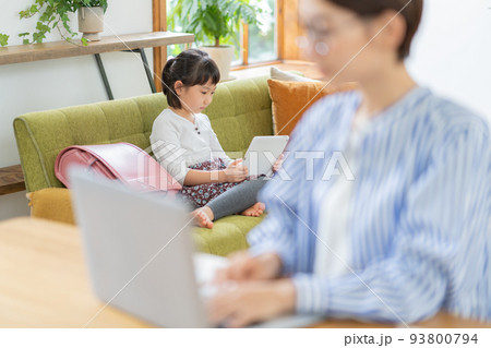 リビングでリモートワークをするママとソファでタブレットを見る小学生の女の子 93800794
