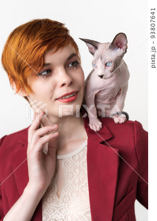 Portrait of hipster redhead young woman with short hair, dressed in red jacket and playful Sphynx Cat sitting on her shoulder. Studio shot on white background. Part of series. 93807641