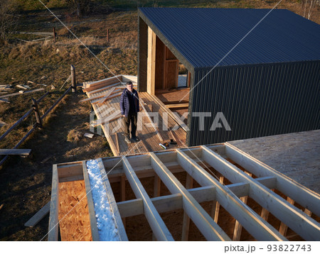 Male architect building wooden frame house in the Scandinavian style barnhouse. Man standing on terrace at construction site, inspecting quality of work in the evening. 93822743