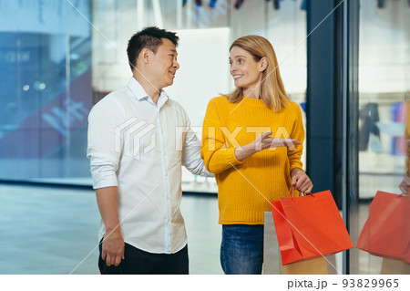 multiracial couple asian man and caucasian woman walking together in mall after shopping with colored bags in hands. walking past shop windows with clothes cheerful family hugging smile. 93829965
