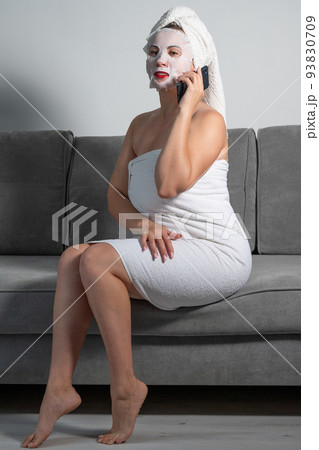 A woman is talking on the phone at home, a woman with a mask on her face and a towel is sitting on the sofa. 93830709