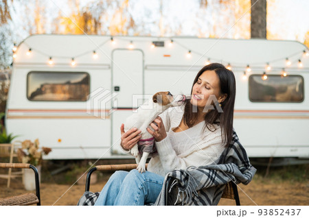 Caucasian woman sitting by a wagon in a wicker chair and hugging with Jack Russell Terrier dog outdoors. Travel in a camper in autumn. Caucasian woman sitting by a wagon in a wicker chair and hugging with Jack Russell Terrier dog outdoors. Travel in a camper in autumn. 93852437