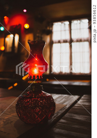Small oil lamp on the table. Old oil lantern with window on background. Red kerosene lamp. Cozy rustic interior. Evening concept. Ancient decorative lamp on table. Vintage home objects. Home decor. 93856612