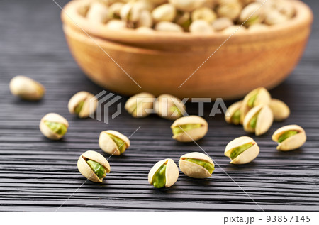 pistachios spill out of a wooden bowl on a black table. 93857145