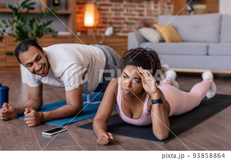 Smiling young black man supports tired sad wife in sportswear do plank exercises on mat 93858864