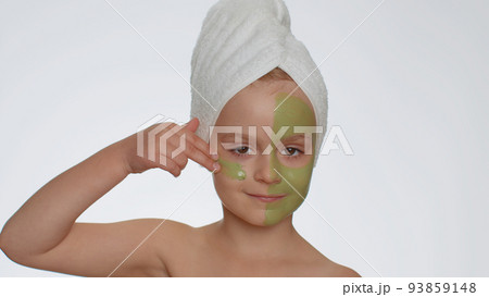 Beautiful smiling child girl kid in towel on head applying cleansing moisturizing green facial mask 93859148