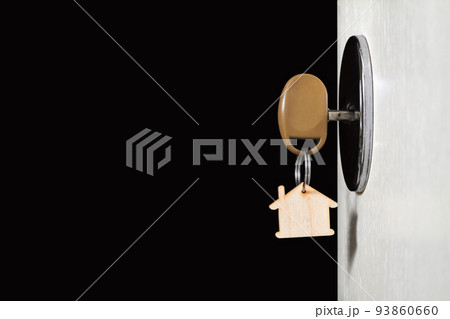 Key with a trinket in the form of a house in the door lock of an open door against the dark background 93860660