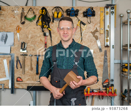 Portrait of a carpenter in goggles and overalls holding a wooden hammer in the workshop against the background of a wall with tools. Portrait of a carpenter in goggles and overalls holding a wooden hammer in the workshop against the background of a wall with tools. 93873268