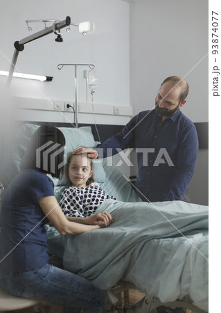 Caring parents standing beside hospitalized sick daughter inside healthcare clinic pediatric ward. Loving mother and father taking care of ill little girl resting in patient bed. Caring parents standing beside hospitalized sick daughter inside healthcare clinic pediatric ward. Loving mother and father taking care of ill little girl resting in patient bed. 93874077