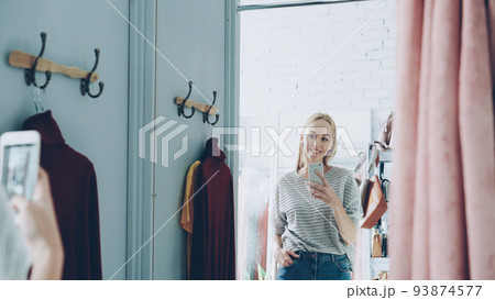 Pretty blond woman is making mirror selfie with smart phone while standing in nice fitting room in luxurious clothes boutique. She is posing, moving and smiling carelessly. 93874577