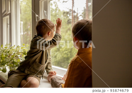 Children sitting on windowsill and waiting for someone comming. Two brothers, friends. Cute preschool kids alone at home. Boys are waiting for their mother or father. Loneliness. Busy parents. 93874679