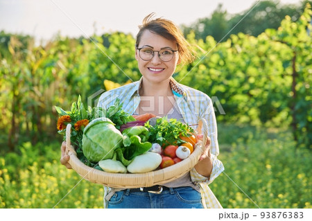 Portrait of smiling woman with basket of different fresh vegetables and herbs Portrait of smiling woman with basket of different fresh vegetables and herbs 93876383