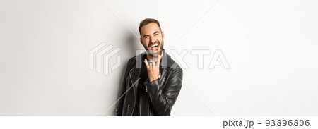 Cool adult man in black leather jacket, showing rock on gesture and tongue, enjoying music festival, standing over white background 93896806