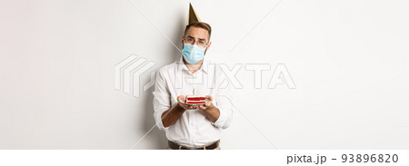 Coronavirus, quarantine and holidays. Sad man cant blow candle off birthday cake, wearing face mask and complaining, standing over white background 93896820