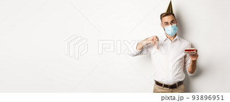 Coronavirus, quarantine and holidays. Man showing thumb down as disappointed with birthday party, wearing face mask and holding bday cake, white background Coronavirus, quarantine and holidays. Man showing thumb down as disappointed with birthday party, wearing face mask and holding bday cake, white background 93896951