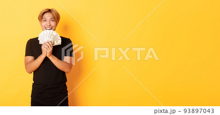 Cheerful winning handsome asian guy, smiling pleased and holding money, feeling lucky, standing yellow background Cheerful winning handsome asian guy, smiling pleased and holding money, feeling lucky, standing yellow background 93897043