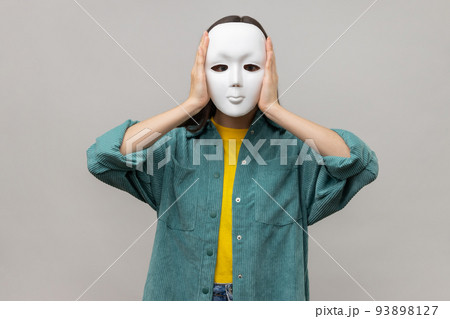 Portrait of unknown woman holding white mask, standing covering face, multiple personality disorder, wearing casual style jacket. Indoor studio shot isolated on gray background. 93898127