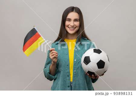Smiling satisfied woman holding in hands flag of Germany and football ball, supporting favourite team on championship, wearing casual style jacket. Indoor studio shot isolated on gray background. Smiling satisfied woman holding in hands flag of Germany and football ball, supporting favourite team on championship, wearing casual style jacket. Indoor studio shot isolated on gray background. 93898131