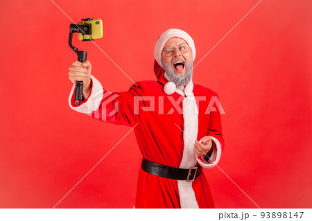 Happy excited elderly man with gray beard wearing santa claus costume with steadicam in hands, livestream, looking at phone screen and screaming. Indoor studio shot isolated on red background. 93898147