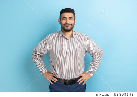 Portrait of happy attractive bearded businessman with smile holding hands on hips, looking at camera with confident expression, wearing striped shirt. Indoor studio shot isolated on blue background. 93898148