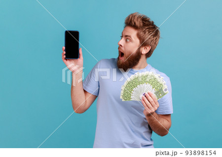 Portrait of astonished amazed bearded man holding euro banknotes and looking at cell phone with empty display, mock up for advertise. Indoor studio shot isolated on blue background. 93898154