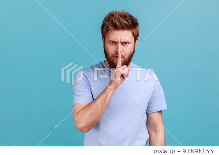 Portrait of bearded man holding finger near lips, showing shh gesture asking to keep silence and be quite, privacy, looking at camera. Indoor studio shot isolated on blue background. Portrait of bearded man holding finger near lips, showing shh gesture asking to keep silence and be quite, privacy, looking at camera. Indoor studio shot isolated on blue background. 93898155