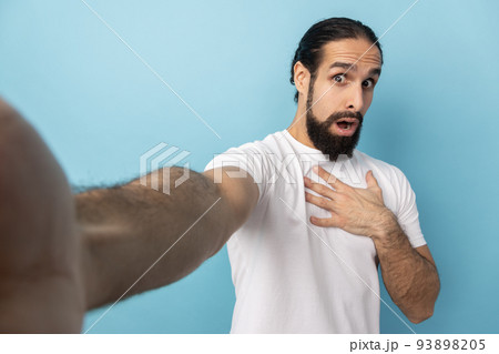 Portrait of scared shocked man with beard wearing white T-shirt looking at camera open mouth, keeps hand on chest, POV, point of view of photo. Indoor studio shot isolated on blue background. Portrait of scared shocked man with beard wearing white T-shirt looking at camera open mouth, keeps hand on chest, POV, point of view of photo. Indoor studio shot isolated on blue background. 93898205