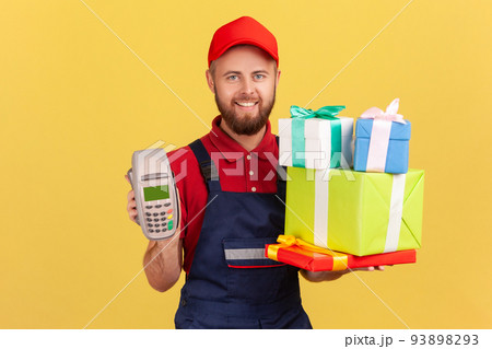 Portrait of positive delighted courier man wearing blue overalls holding stack of present boxes and pos terminal for online paying. Indoor studio shot isolated on yellow background. 93898293