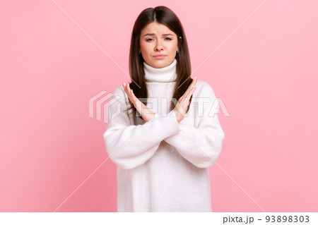 Portrait of determined brunette young woman showing x sign with crossed hands, stop, this is the end, wearing white casual style sweater. Indoor studio shot isolated on pink background. 93898303