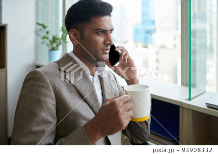 Portrait of serious Indian entrepreneur drinking morning coffee and looking through office window 93908332