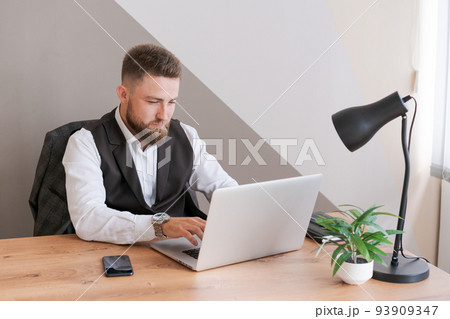 Caucasian man sits at table near laptop in office and looks at screen. Serious Caucasian man sits at table near laptop in office and looks at screen. Serious 93909347