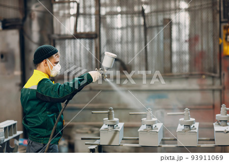 Powder primer coating of metal parts. Worker man in a protective suit sprays powder paint from gun on metal product construction at factory plant. 93911069