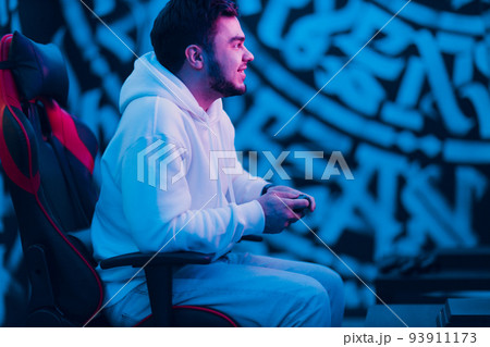 Young man plays on game console with gamepad sitting on chair. Young man plays on game console with gamepad sitting on chair. 93911173