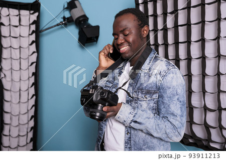 Photographer talking on smartphone while standing on blue background. Young producer having a phone call while working on production studio equipped with professional softboxes and spotlights. Photographer talking on smartphone while standing on blue background. Young producer having a phone call while working on production studio equipped with professional softboxes and spotlights. 93911213