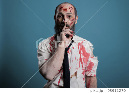 Wounded corpse doing hush silence sign in front of studio camera, showing mute shh gesture about confidential secret. Cruel dangerous zombie acting creepy and spooky, sinister demon. 93911270